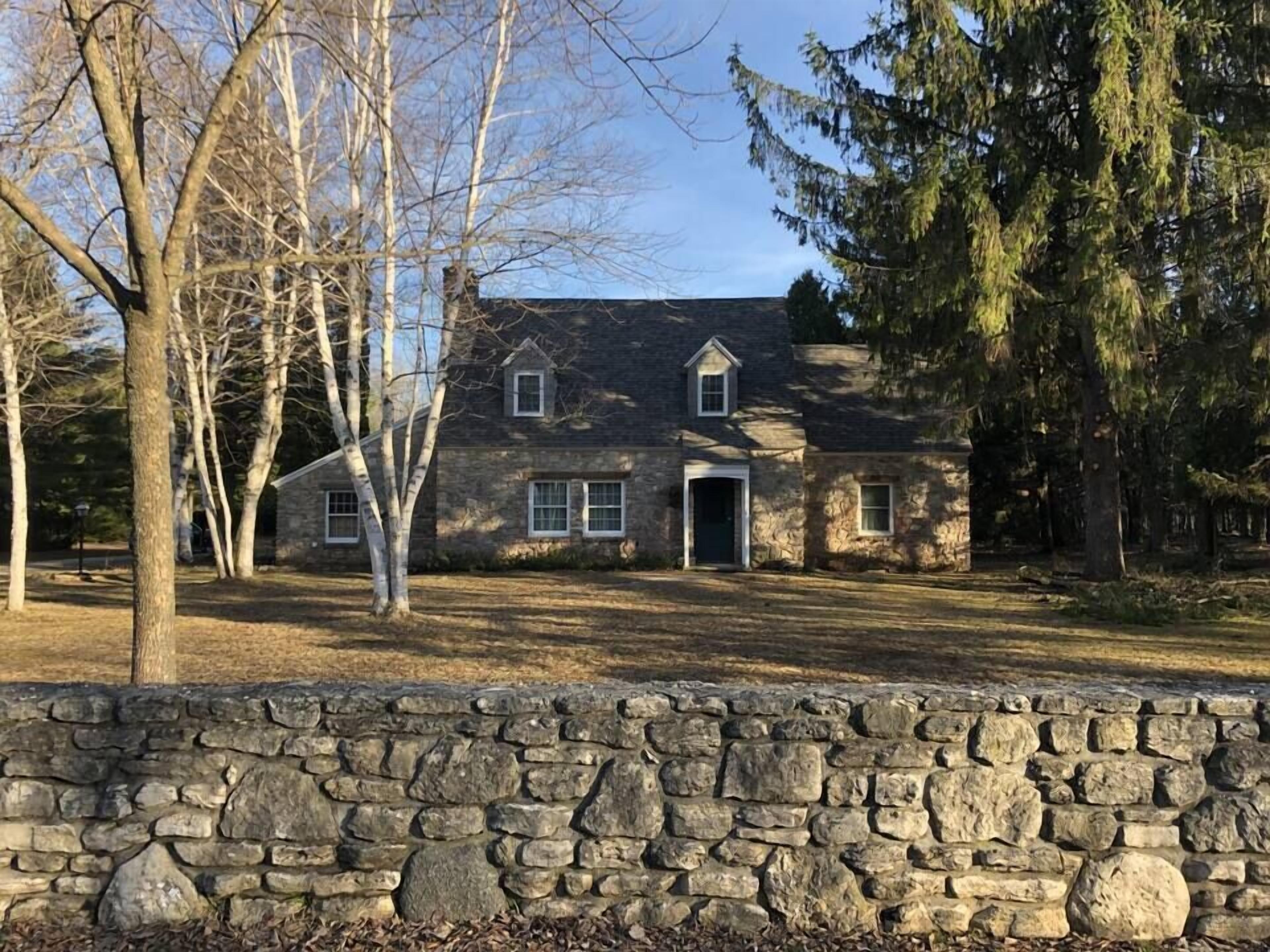 A stone house with a stone wall in front, surrounded by trees.