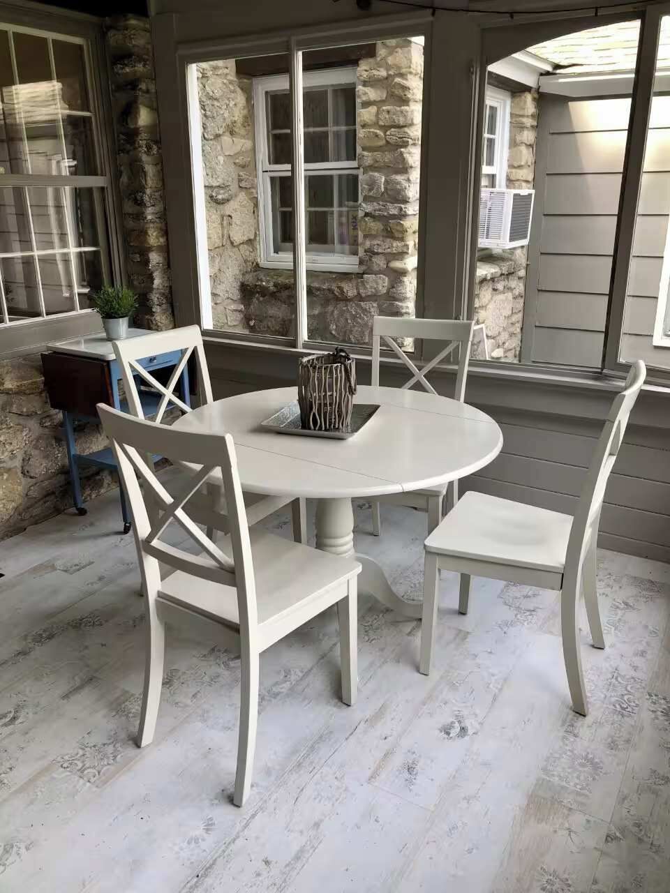 Een ronde tafel met vier witte stoelen, een stenen muur en een raam met zonwering.