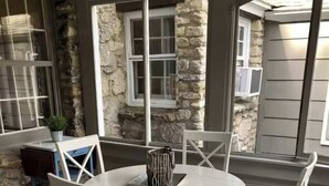 Una mesa redonda con cuatro sillas blancas, un muro de piedra y una ventana con persianas.
