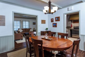 A dining area with a wooden table, chairs, a chandelier, and a kitchen in the background. - Manistique Stone Cottage - Next to Peninsula State Park (Fish Creek)