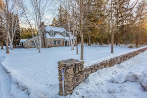 A stone house with a snow-covered roof, surrounded by trees and a stone wall.