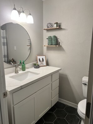Een badkamer met een wit wastafelmeubel, een ronde spiegel, een wastafel en een toilet. Boven het wastafelmeubel zijn planken met toiletartikelen en een lijst met een foto.