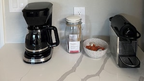 Una encimera de cocina con una cafetera, un tarro de café, un bol de granos de café y un cartel con la carta de café.