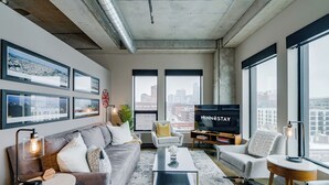 Uma sala de estar moderna com uma TV grande, assentos confortáveis, fotografias emolduradas da paisagem urbana e amplas janelas com vista para a cidade.