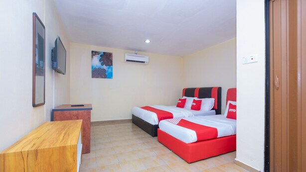 A hotel room with a wooden desk, two beds with red and white bedding, a small table, a wall-mounted air conditioner, and a painting. - OYO 89921 Motel New Bay Sg. Lembing (Sungai Lembing)