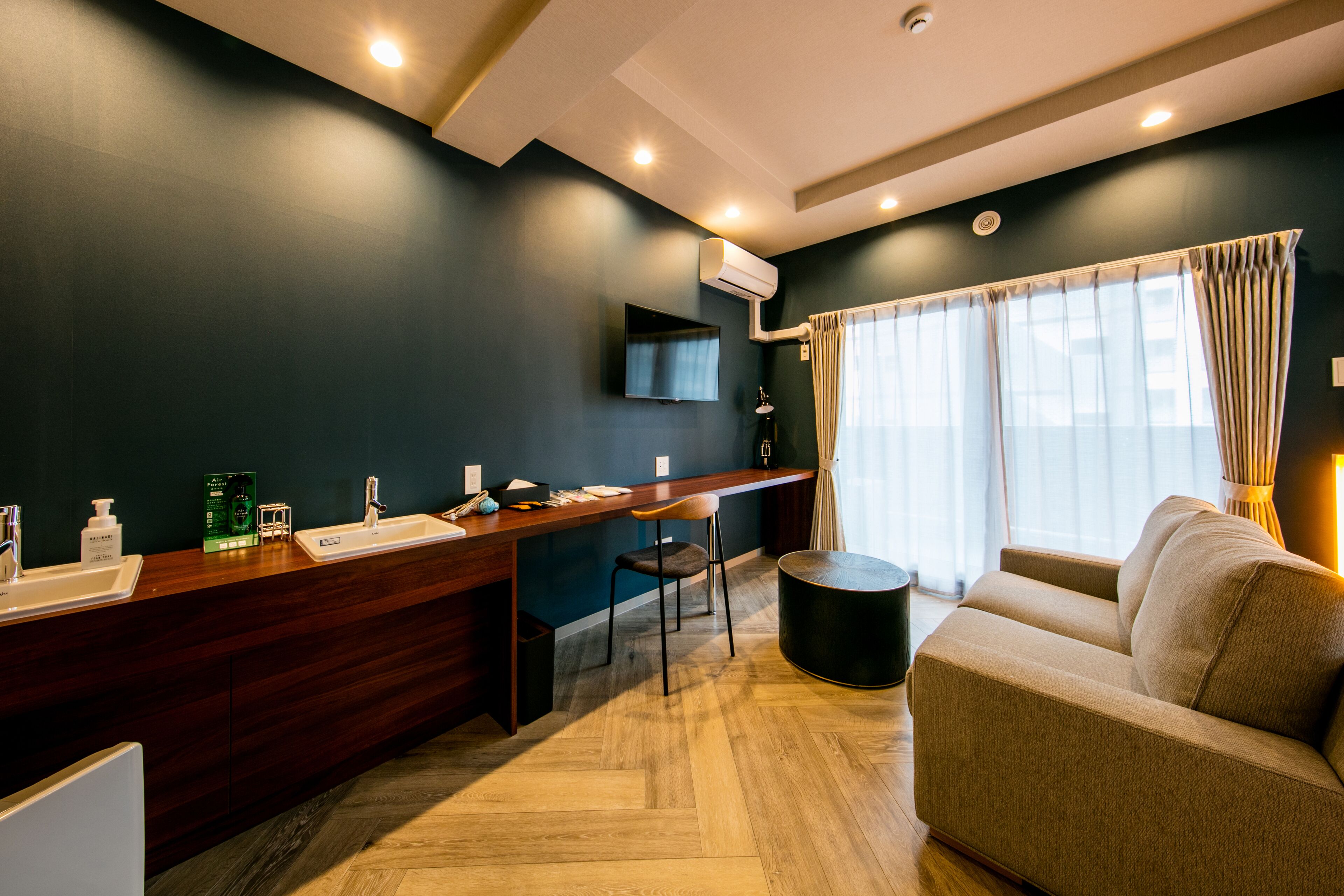 A modern hotel room with a dark blue accent wall, a wooden desk with a chair, a sofa, and a large window with curtains.
