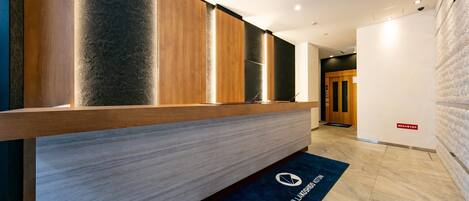 A reception area with a wooden reception desk, a dark blue mat with the hotel logo, and a modern interior design.