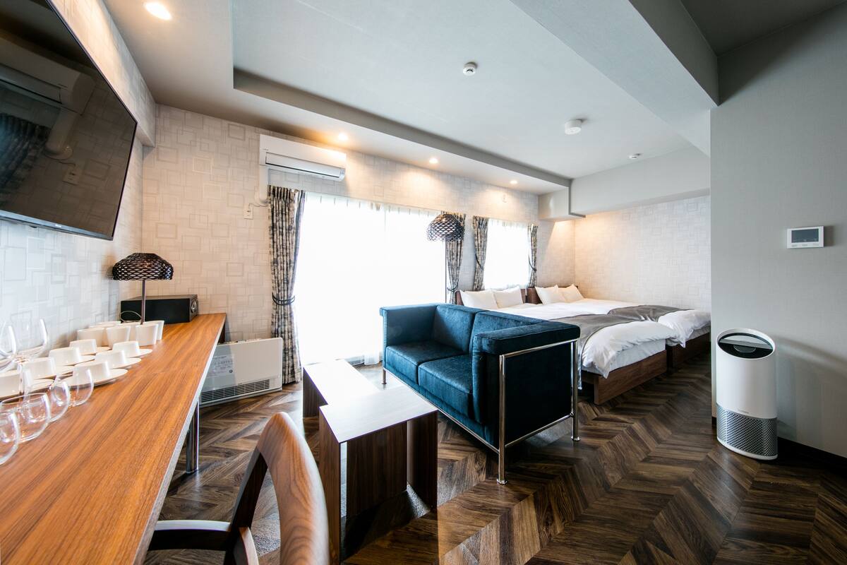 A modern hotel room with a wooden dining table, a blue sofa, a bed, and a white trash bin.