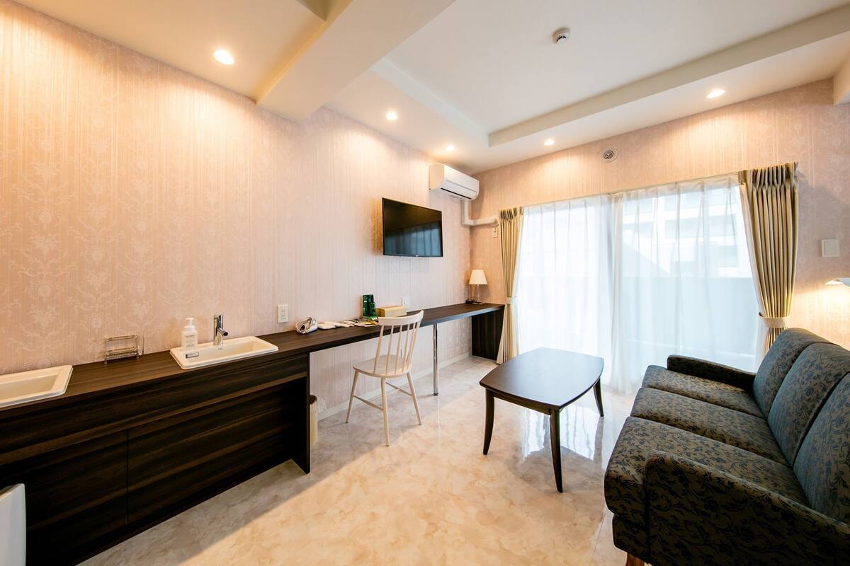 A hotel room with a dark wood vanity, a flat-screen TV, a desk with a chair, a sofa, and a large window with curtains.