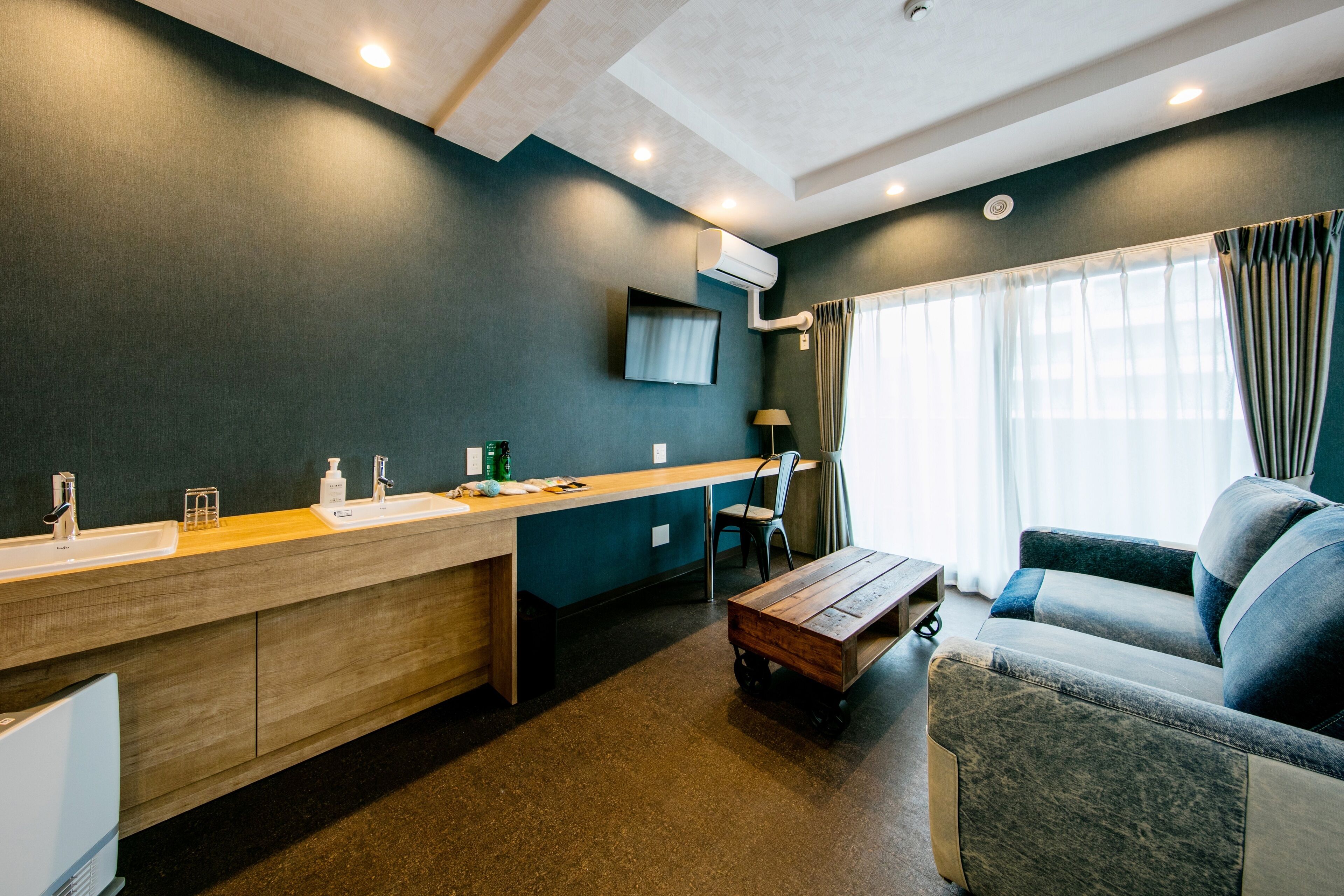 A modern hotel room with a kitchenette, a sofa, a wooden coffee table, and a TV mounted on the wall.