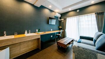 A modern hotel room with a kitchenette, a sofa, a wooden coffee table, and a TV mounted on the wall.