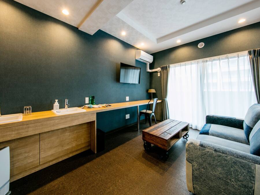 A modern hotel room with a kitchenette, a sofa, a wooden coffee table, and a TV mounted on the wall.