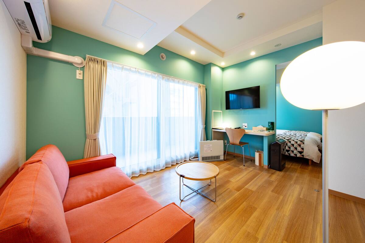 A modern room with a teal wall, a wooden floor, a red sofa, a small round table, a desk with a chair, a bed with a checkered blanket, and a mounted TV.