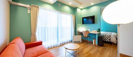 A modern room with a teal wall, a wooden floor, a red sofa, a small round table, a desk with a chair, a bed with a checkered blanket, and a mounted TV.