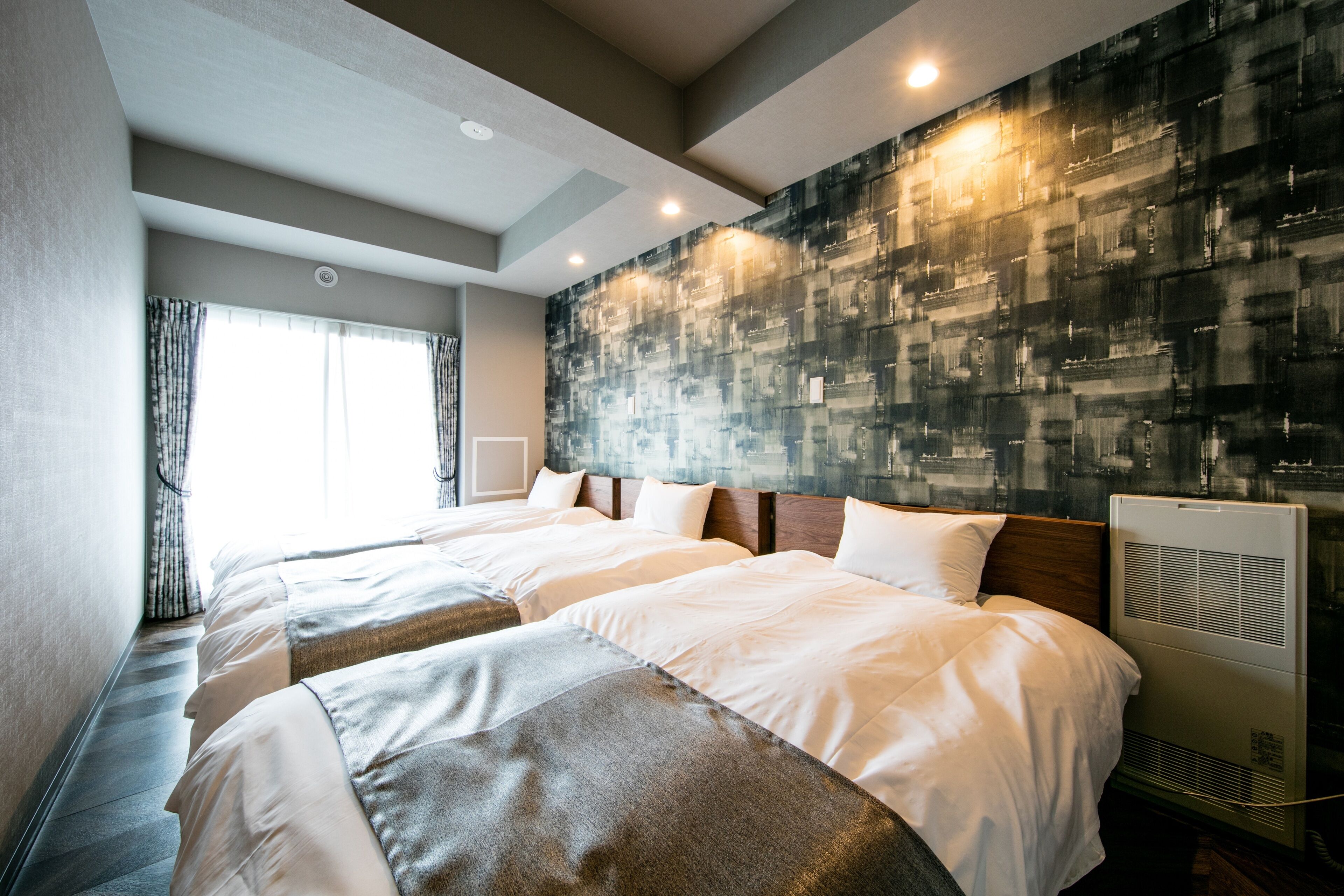 Habitación de hotel con dos camas, un ventanal con cortinas, una pared con textura y un techo con iluminación empotrada.