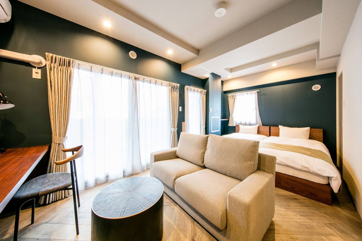 A modern hotel room with a sofa, a round coffee table, a wooden desk, and a bed with a wooden headboard.