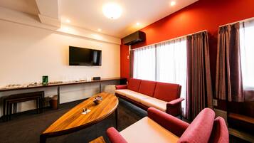 Habitación de hotel moderna con una mesa de comedor de madera, un sofá rojo, un televisor de pantalla plana en la pared y una ventana con cortinas.