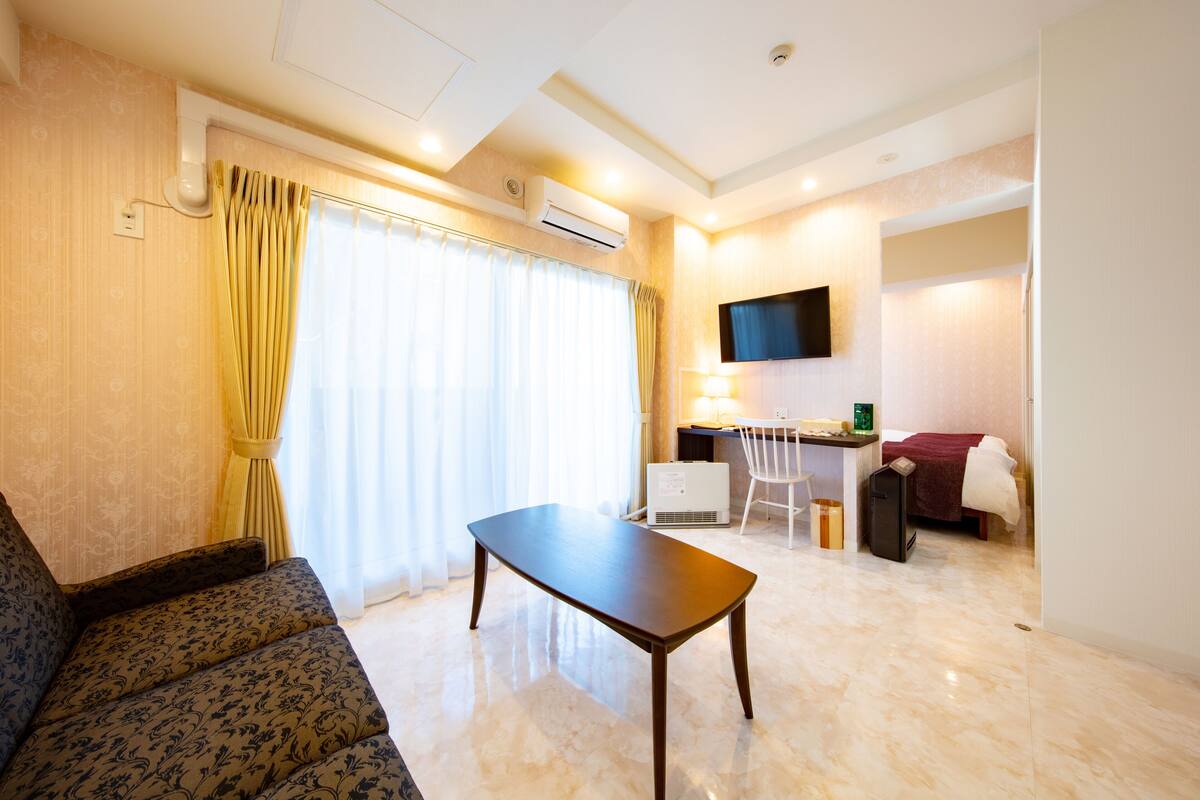 A hotel room with a sofa, a table, a desk with a chair, a TV mounted on the wall, and an air conditioning unit.