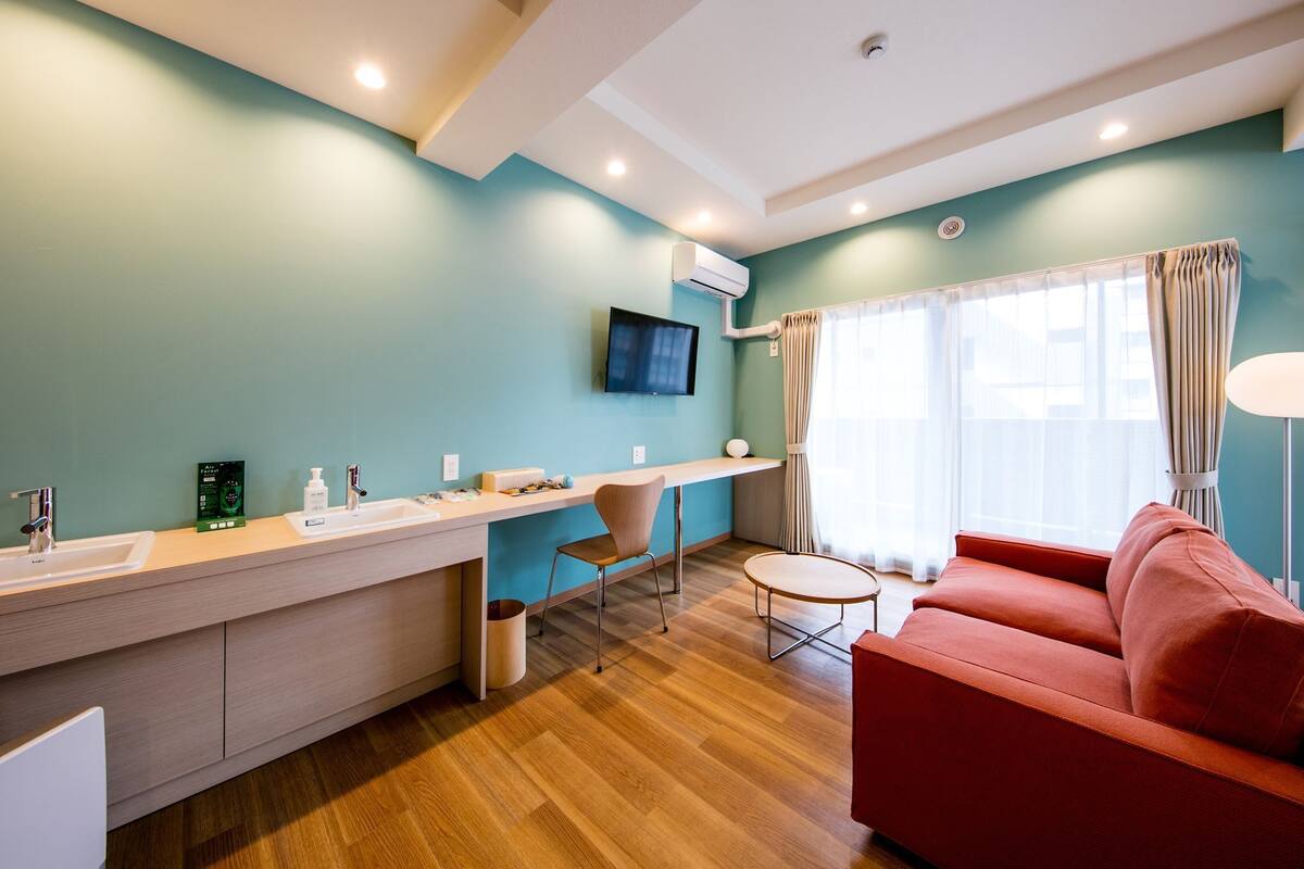 A modern room with a sofa, a small round table, a TV mounted on the wall, a window with curtains, and a built-in sink.