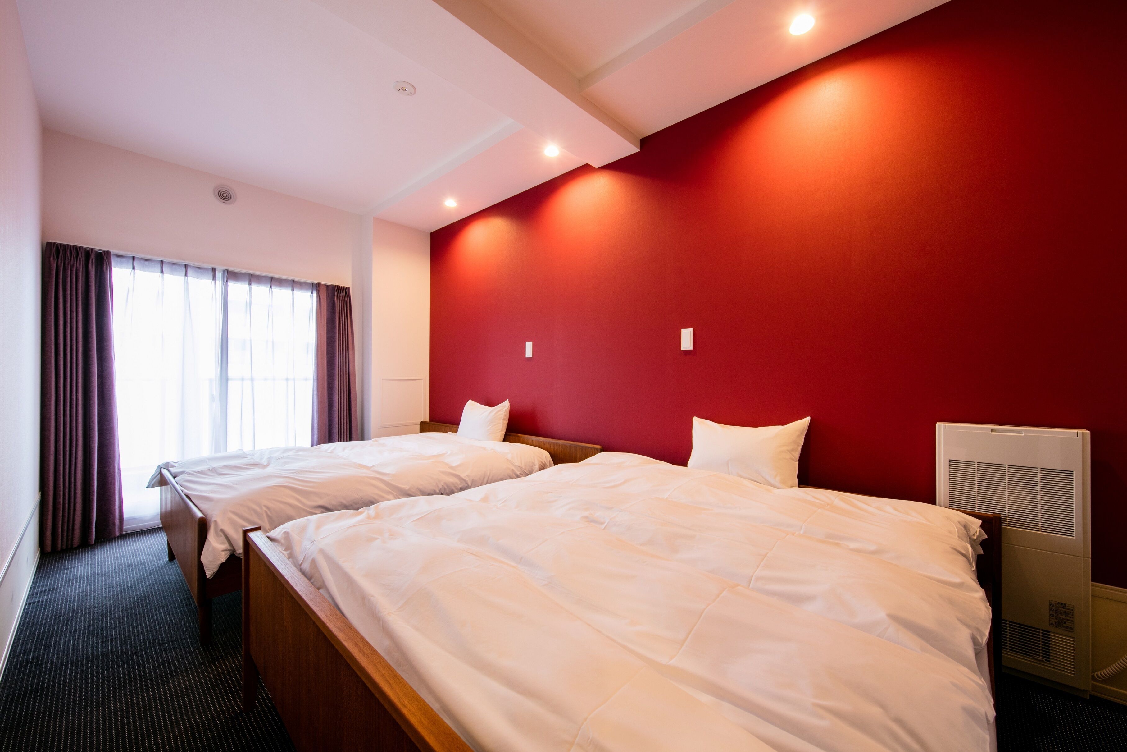 A hotel room with two beds, a red accent wall, and a window with curtains.