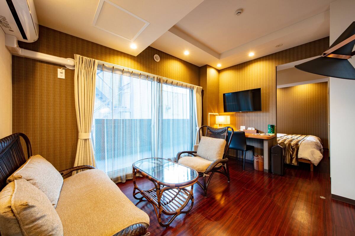 A hotel room with a wooden floor, a glass table, two wicker chairs, a bed, a desk with a chair, a TV mounted on the wall, and a window with curtains.