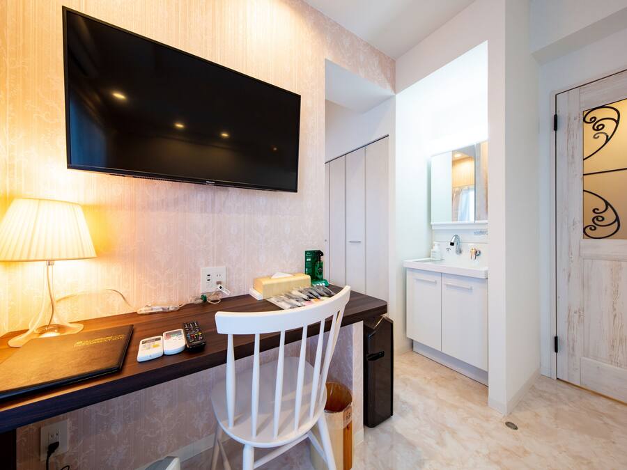 A hotel room with a desk, a chair, a television mounted on the wall, a lamp, a small refrigerator, and a trash bin.