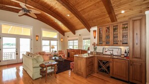 A spacious living area with a wooden ceiling, a large window, and a kitchen with wooden cabinets and a wine rack.