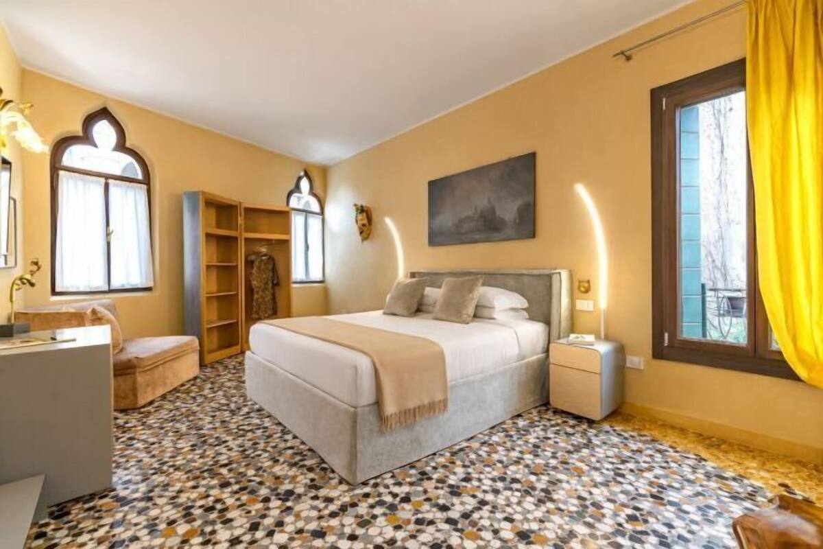 Ein Schlafzimmer mit Bett, Nachttischen, Schreibtisch, Stuhl, einem Fenster mit Vorhängen und einem gemusterten Boden.