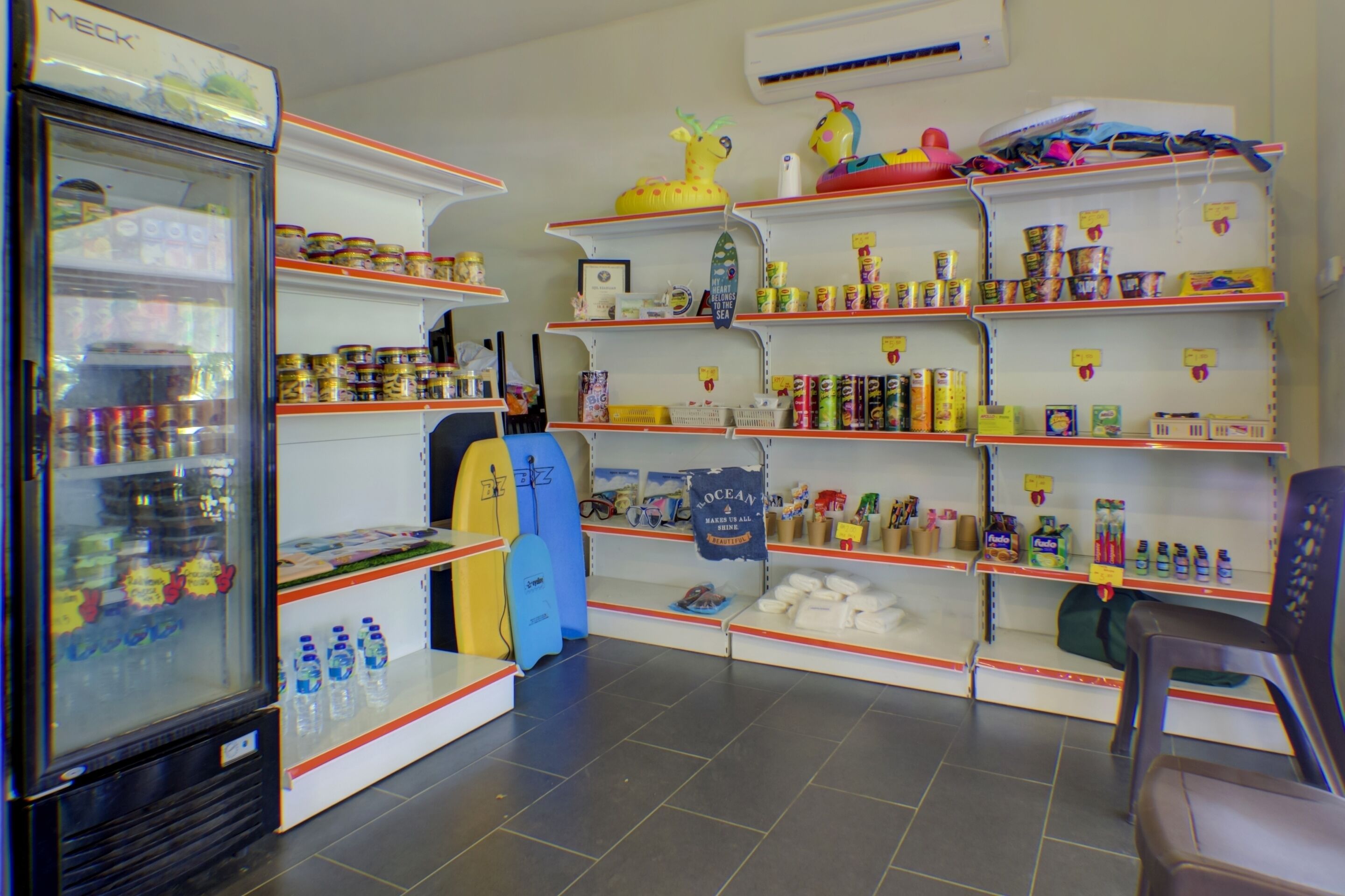 Una tienda con estantes llenos de diversos productos, un refrigerador y una tabla de surf.