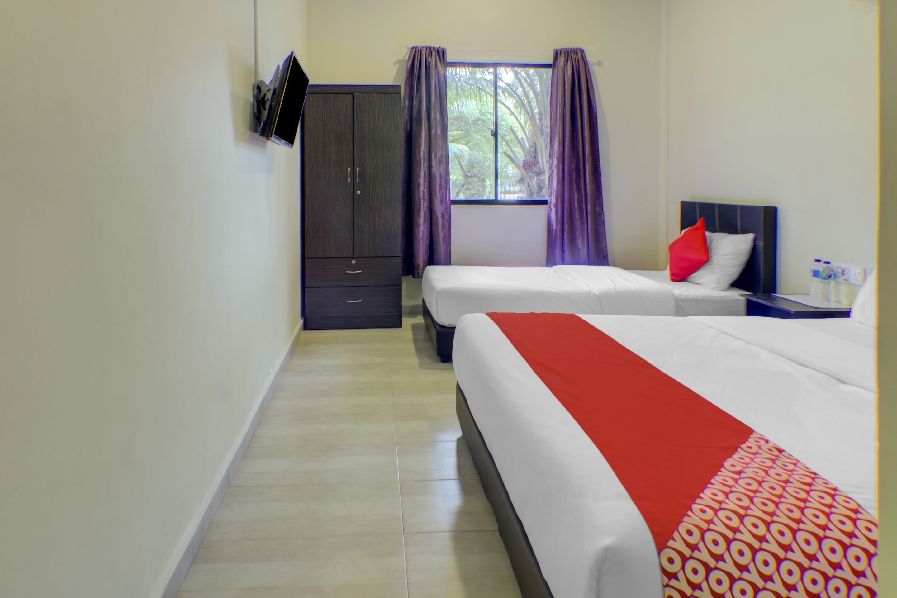A hotel room with two beds, a wardrobe, a TV mounted on the wall, and a window with curtains.