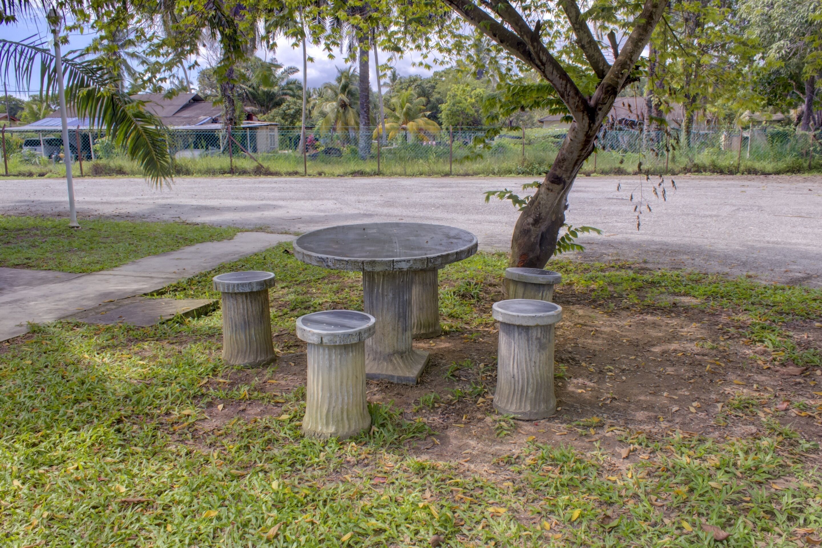 Un espace extérieur aménagé avec une surface en béton, comprenant une table ronde et quatre tabourets, le tout entouré de pelouse et d’arbres.