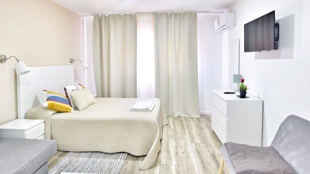 Une chambre d’hôtel moderne équipée d’un lit, d’une table de chevet, d’une lampe, d’une télévision et d’un petit bureau agrémenté d’une plante.