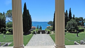 A view through columns to a paved pathway leading to a building with a red-tiled roof, surrounded by greenery and cypress trees, overlooking a body of water. - Holiday Home Aspalathos, 2 BDR Apt, Close To All Tourist Attractions 		 (Split)