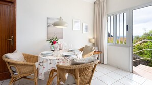 Un coin repas avec des chaises en osier, une table dressée avec des assiettes, des verres et un vase de fleurs. Une fenêtre avec des rideaux offre une vue sur la verdure à l’extérieur.