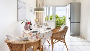 Un coin repas avec des chaises en osier, une table ronde recouverte d’une nappe à motifs floraux et une vue sur l’extérieur, visible à travers une porte vitrée.