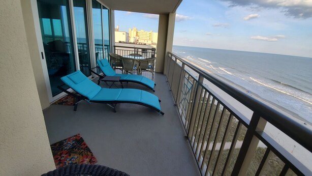 A balcony with two blue lounge chairs, a wicker chair, and a glass door leading to an indoor area.