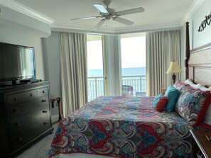 A hotel room with a bed, a TV, a ceiling fan, a balcony with a view of the sea, and a dresser.