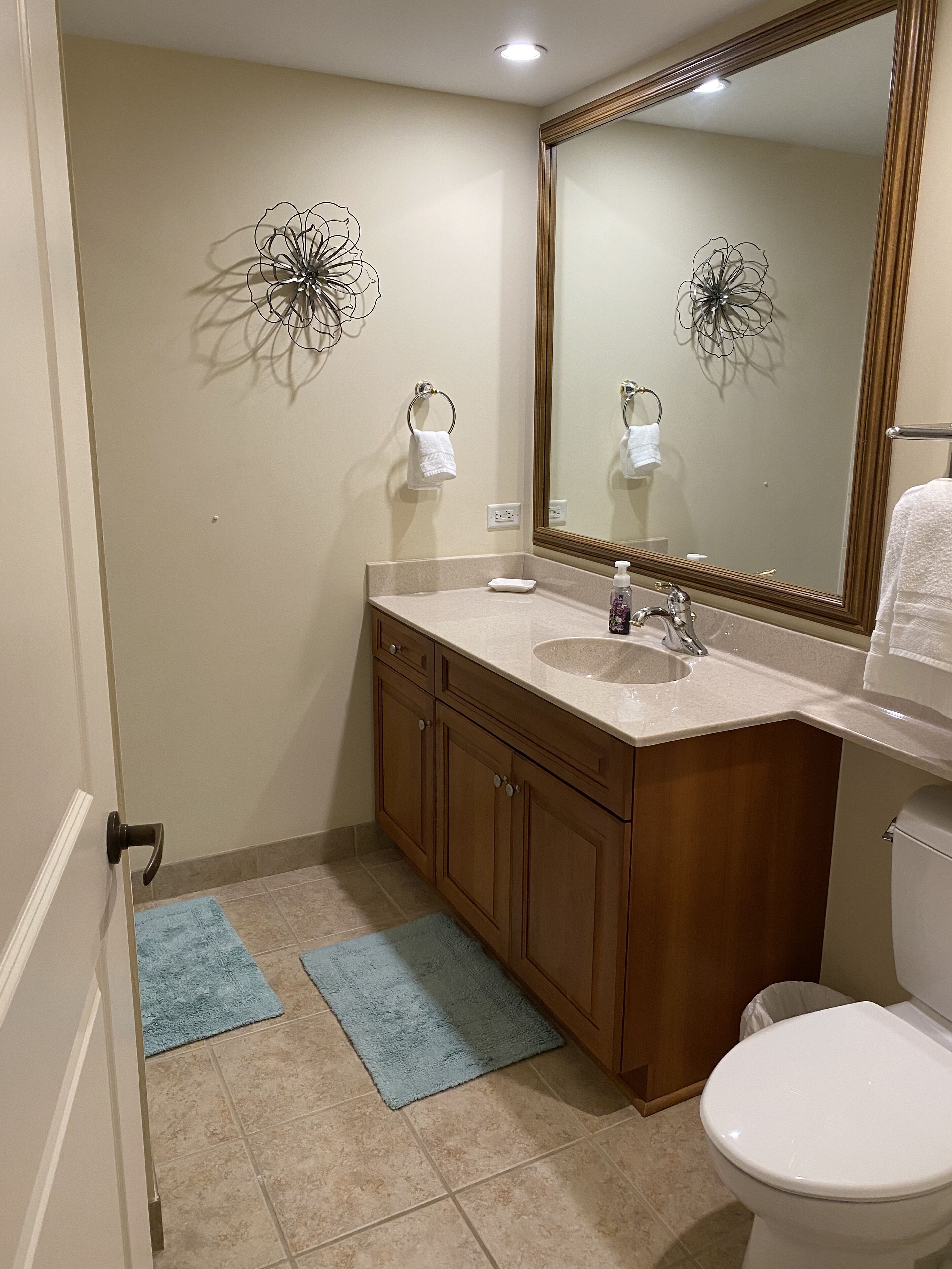 Een badkamer met een beige muur, een houten wastafelmeubel, een witte wastafel, een spiegel en een toilet. Er liggen twee blauwe handdoeken op de vloer.