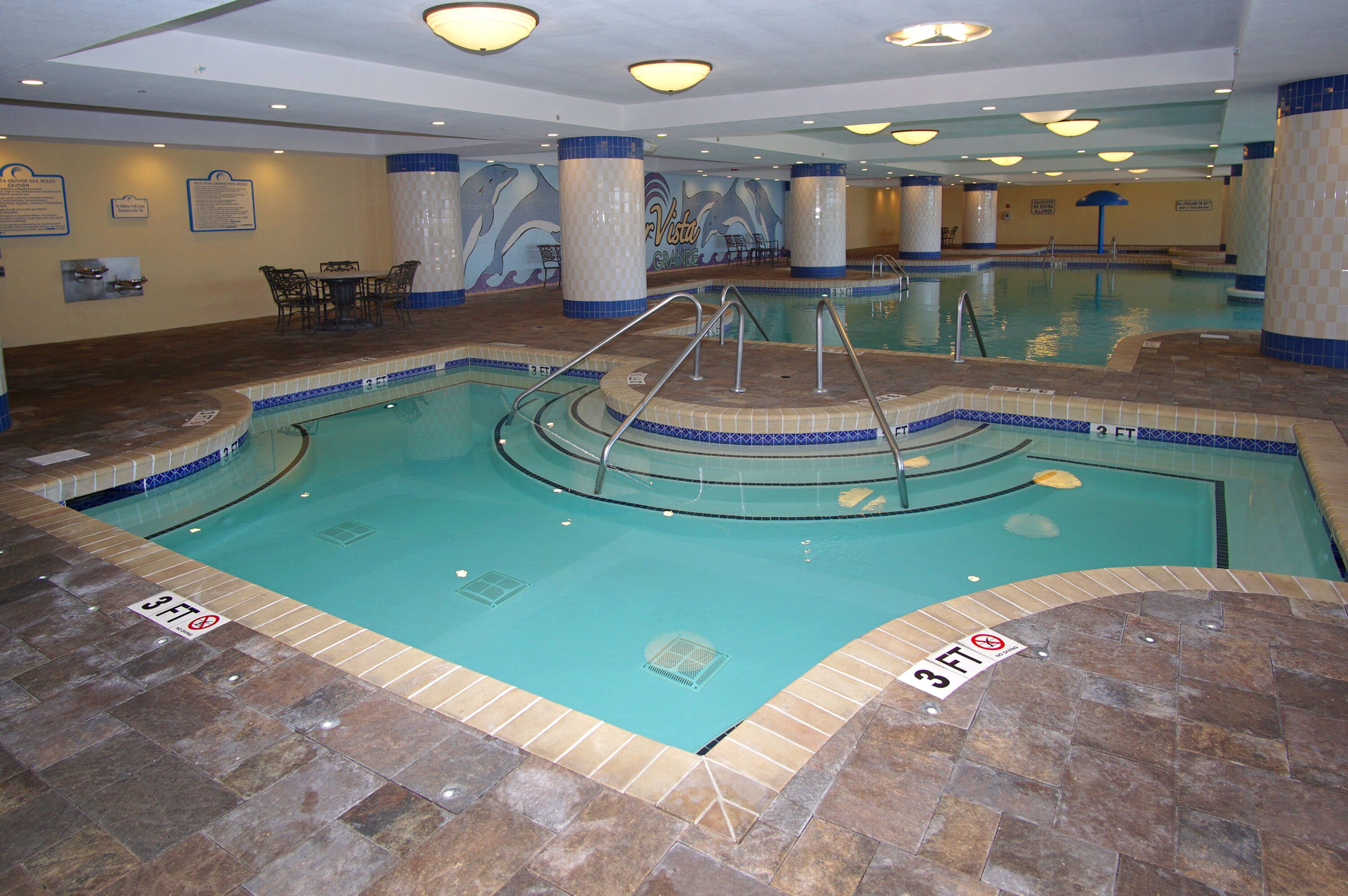 Una piscina interior con indicador de profundidad de 0.9 metros, paredes con azulejos y una zona de estar con mesas y sillas.