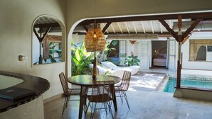 Uma área de refeições com uma mesa redonda e cadeiras de vime, um lustre pendurado e vista para uma piscina e área externa com assentos.