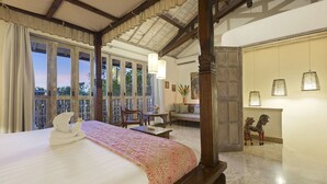 Um quarto espaçoso com uma cama grande, um banco de madeira, uma mesa pequena e uma cadeira. Há janelas amplas com cortinas, uma varanda com guarda-corpo de madeira e vista para árvores.