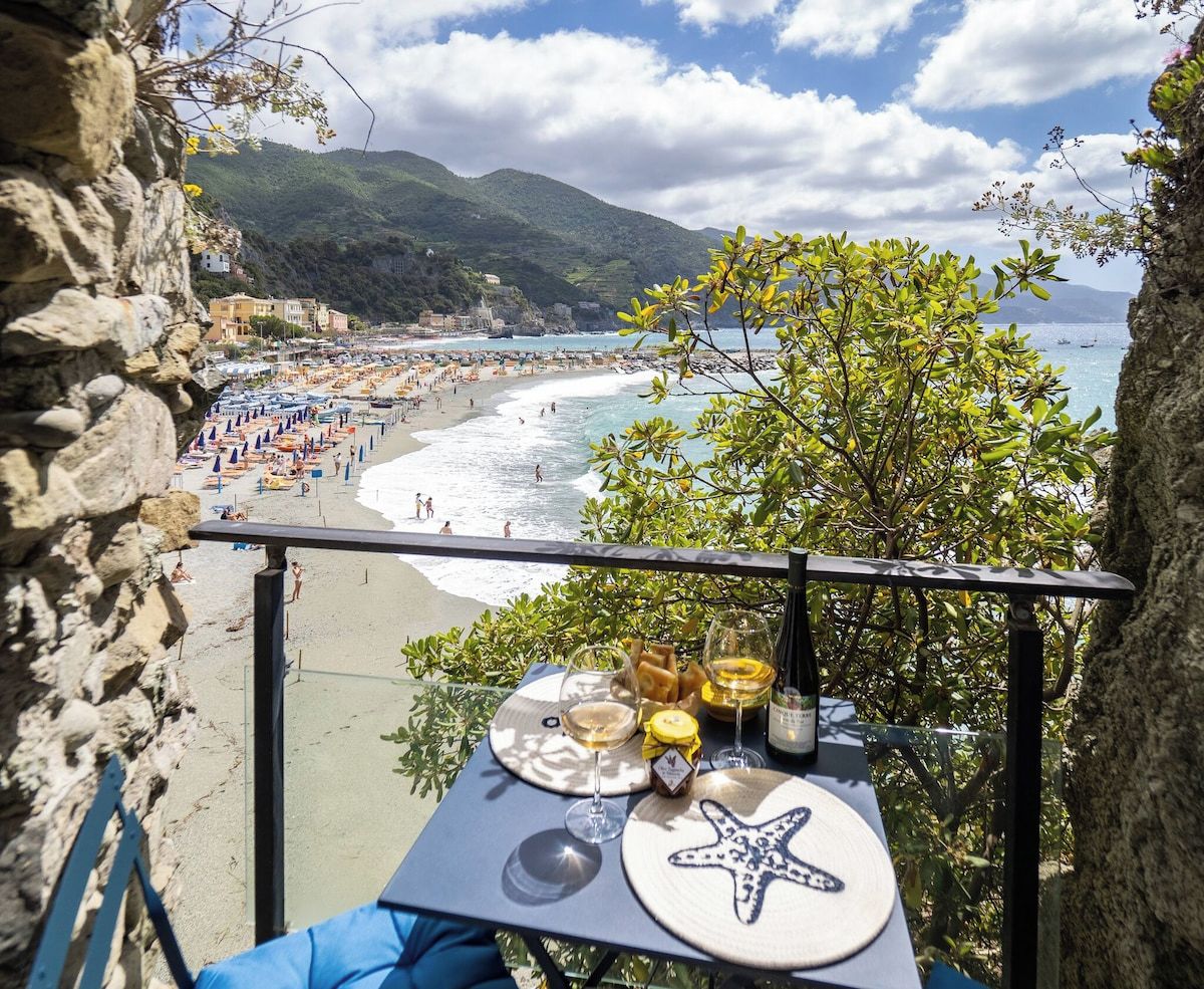 Une table avec un set de table orné d’une étoile de mer, des verres à vin et une assiette de nourriture, le tout donnant sur une plage avec des parasols et des gens.
