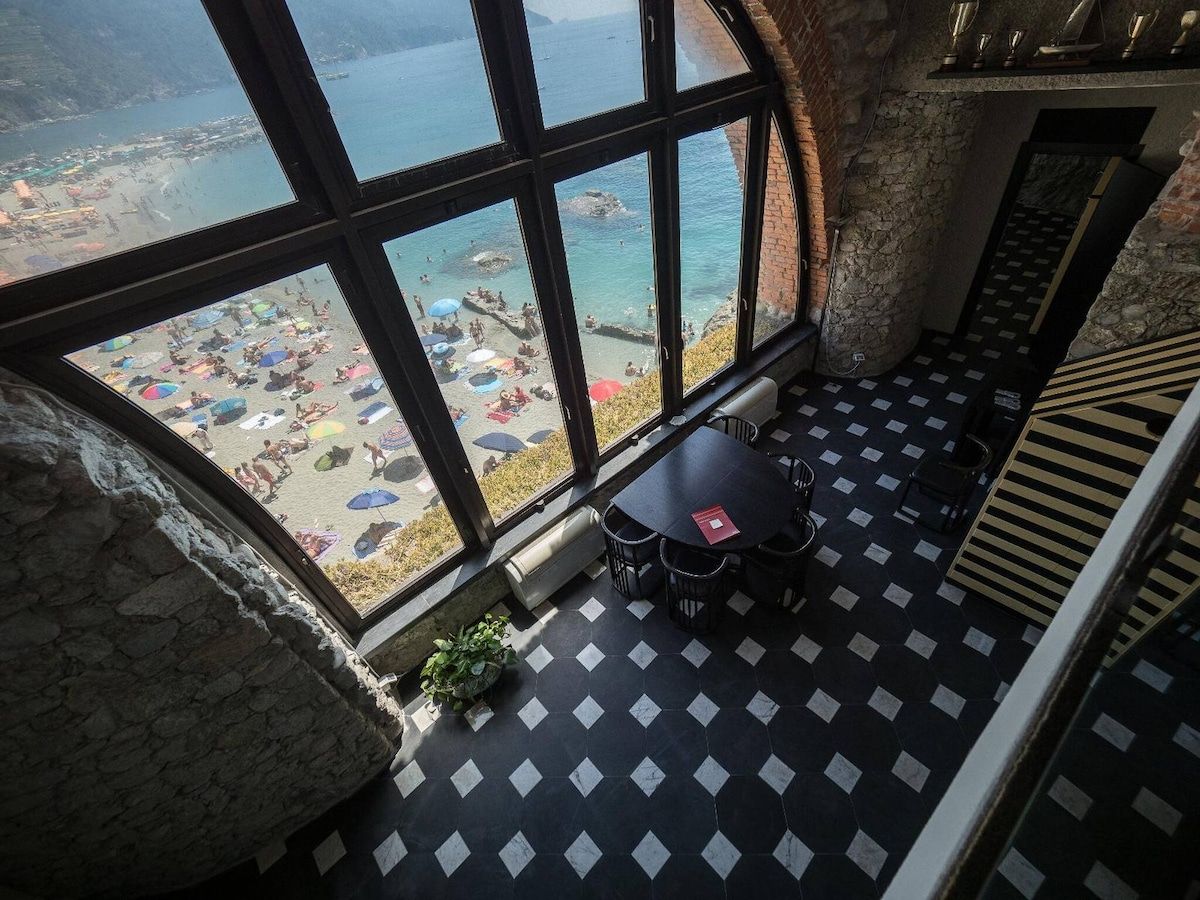 Ein Zimmer mit großem Fenster und Blick auf einen Strand, mit Fliesenboden und einem Tisch mit Stühlen.