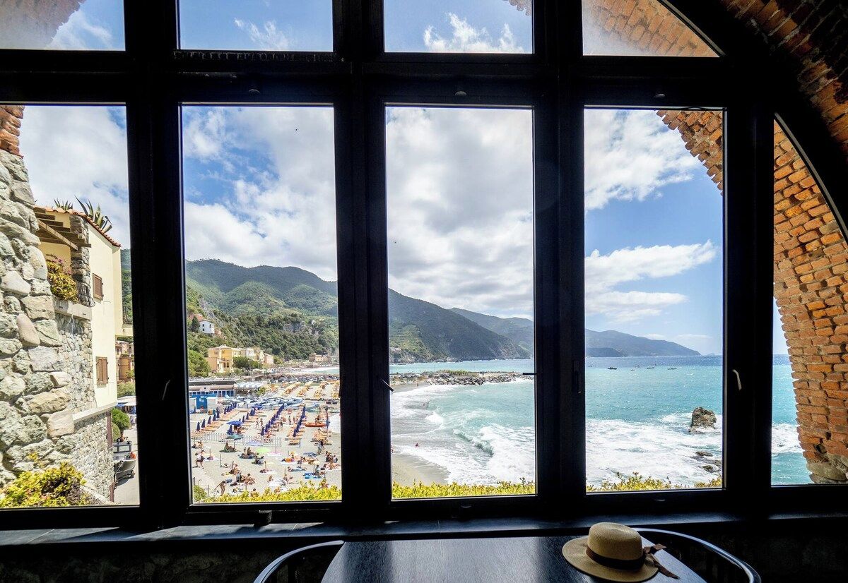 A view from a window overlooking a beach with a clear sky, a rocky coastline, and a building with a stone facade.