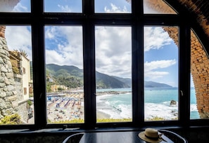 A view from a window overlooking a beach with a clear sky, a rocky coastline, and a building with a stone facade.