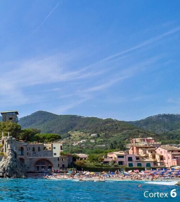 A coastal town with buildings near the water, a beach with umbrellas, and a mountainous backdrop.