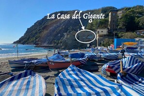 Ein Strand mit gestreiften Sonnenschirmen und Booten, ein Gebäude mit der Aufschrift 'La Casa del Gigante' und einem Bergpanorama im Hintergrund.