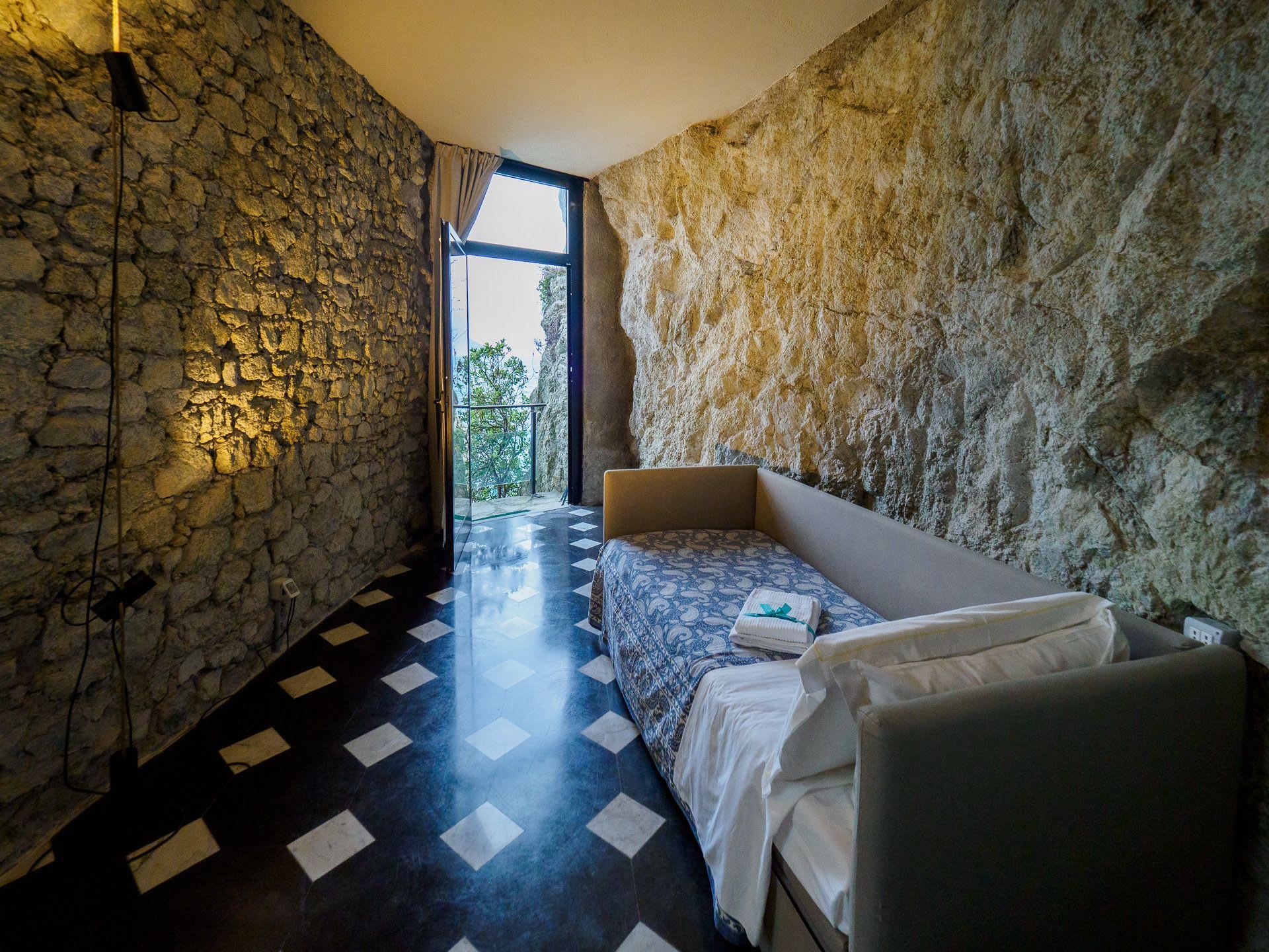 A room with a stone wall, a bed with a patterned bedspread, and a view through a window.