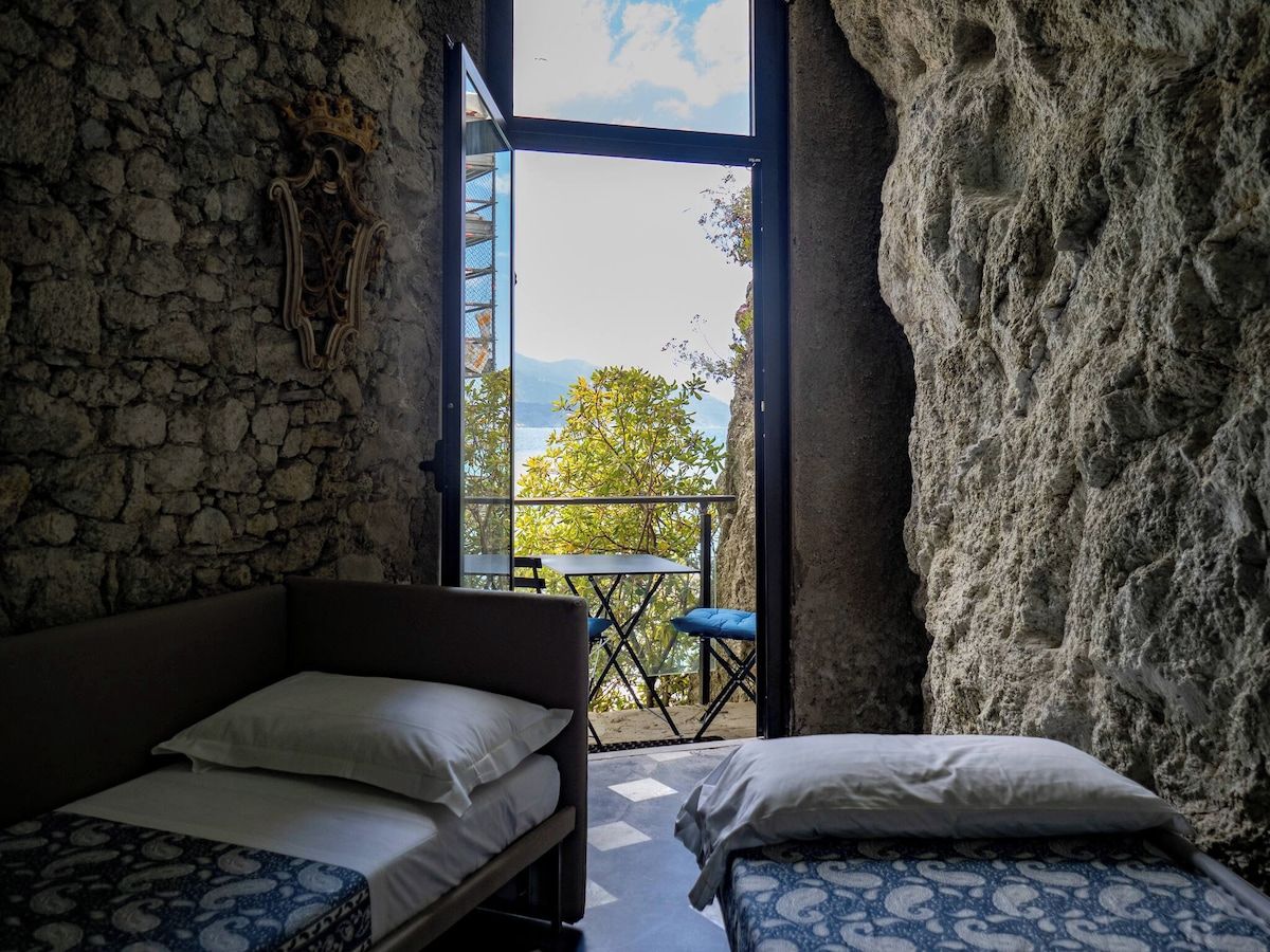 Ein Zimmer mit Steinwand, einem Bett, einem kleinen Tisch und Blick ins Freie.