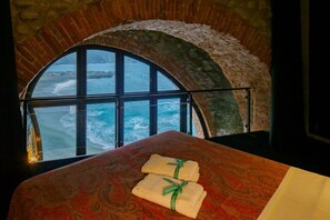 A room with a large arched window overlooking the sea, a bed with a patterned bedspread, and two towels on the bed.